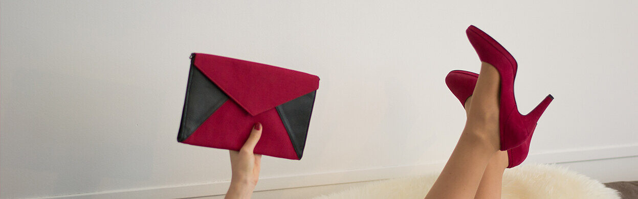 Main tenant une pochette enveloppe framboise et noire à côté de jambes en escarpins rouges